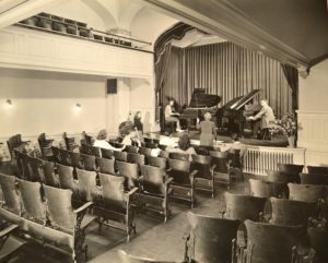 In this 1944 photo of MacDowell Hall, the pipe organ console may be seen in the front left corner. Turned balusters have replaced the former solid wall of the proscenium. Miss Esther Bienfang, who taught piano for many years, is giving instruction to two of her students. Whitman Archives.